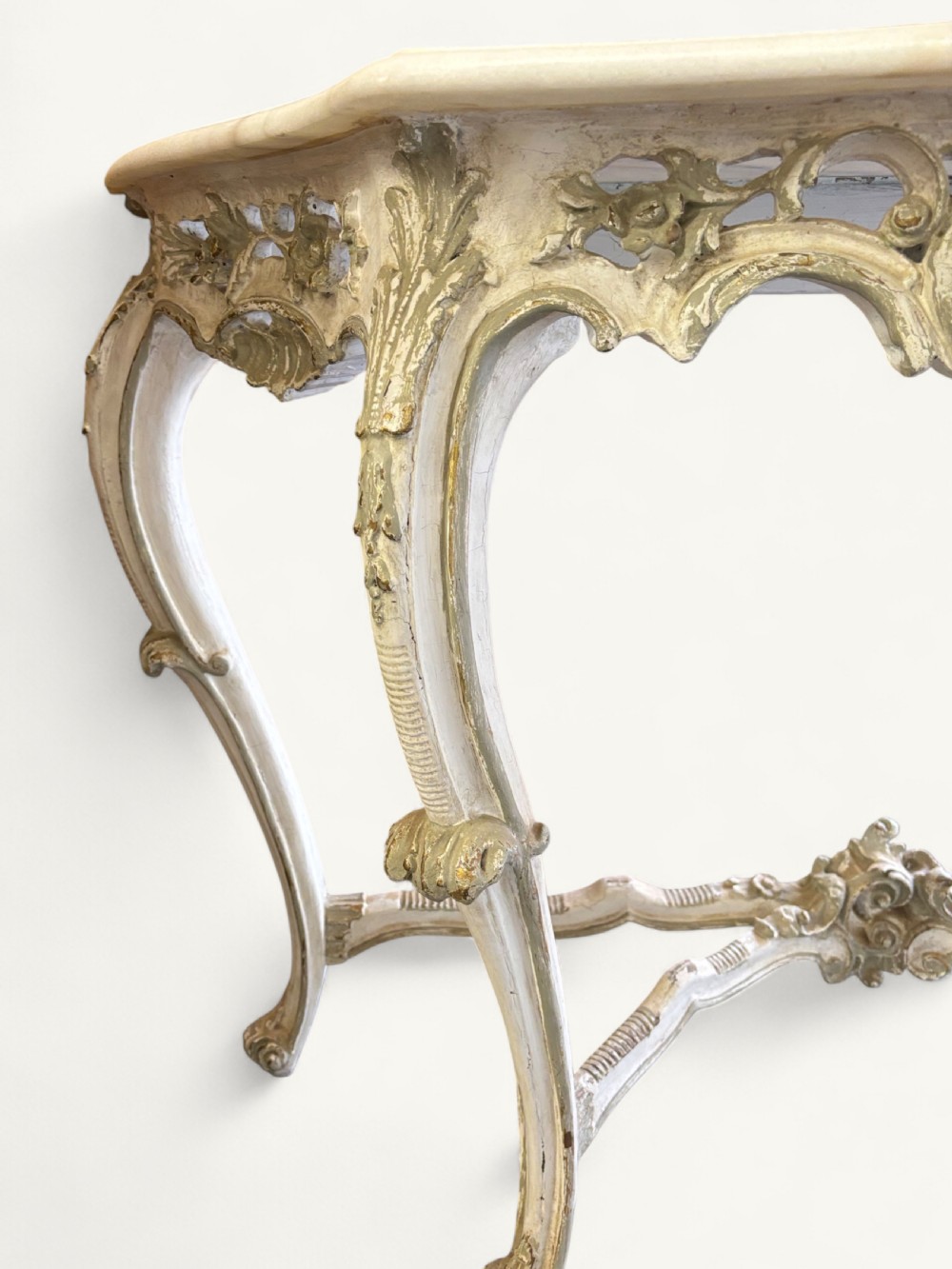A French Rococo style console table featuring a light marble top, off-white distressed paint finish, cabriole legs, and an intricately carved apron with scrolls. View XI