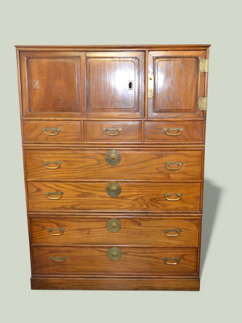 A tall Taisho period Japanese elm tansu chest featuring prominent wood grain, circular brass escutcheons, sliding top cabinet doors, and multiple drawers.