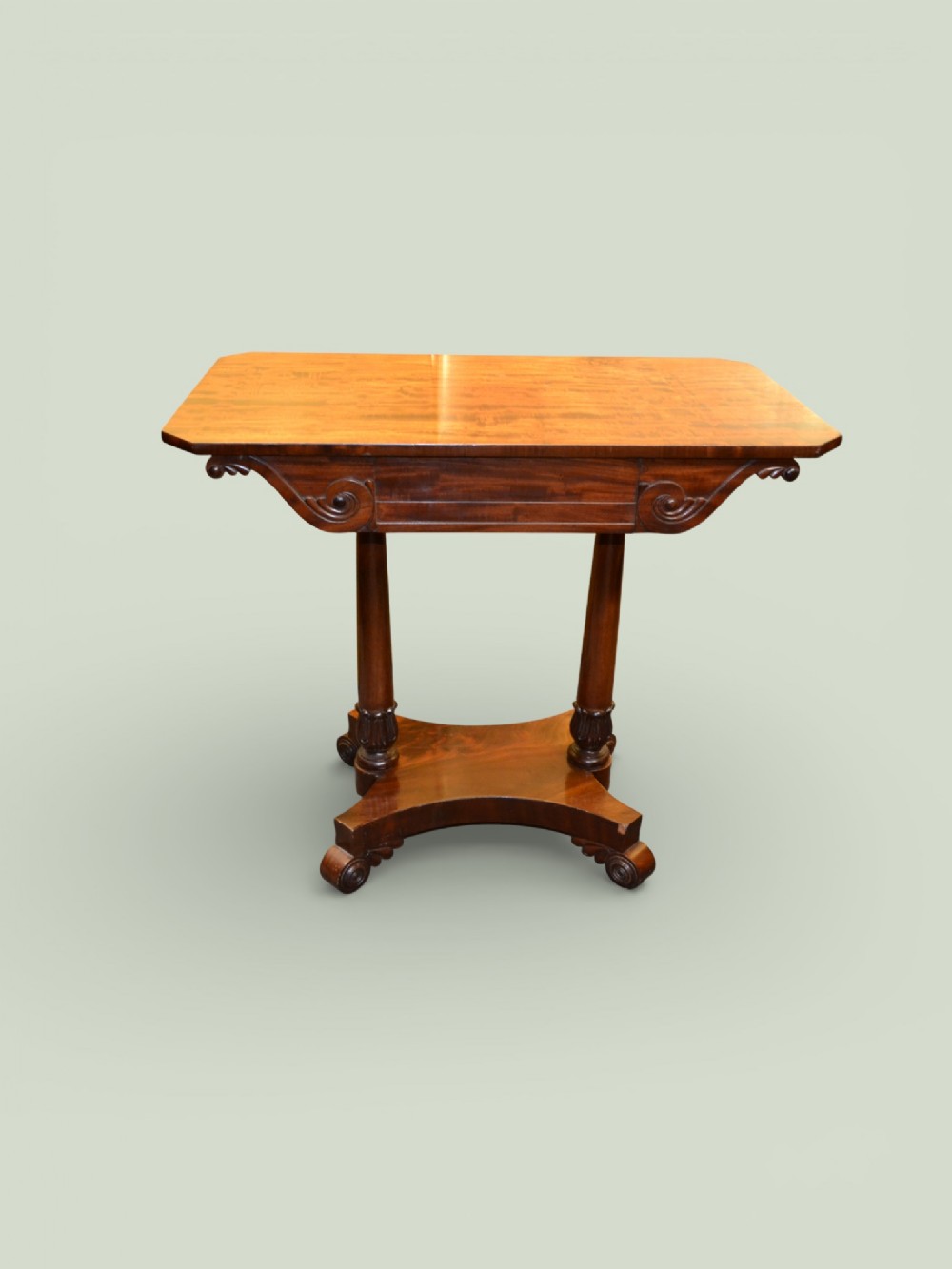 A William IV mahogany side table featuring a rectangular top, frieze drawer with scroll carved brackets, twin column supports, and a shaped plinth base on scroll feet.