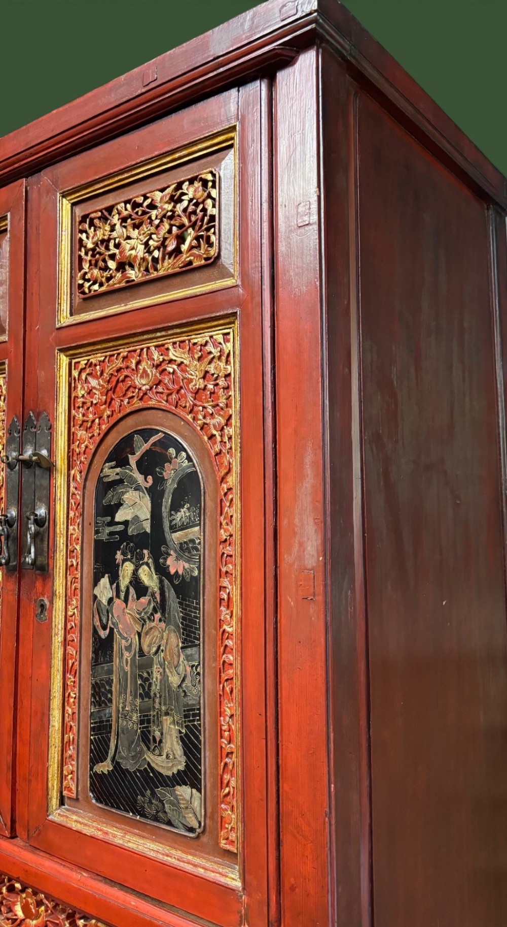 An antique Chinese tall wooden cabinet featuring red lacquered framing, gilded openwork wood carvings, black panels decorated with gold figures and floral motifs, and central brass hardware. View II