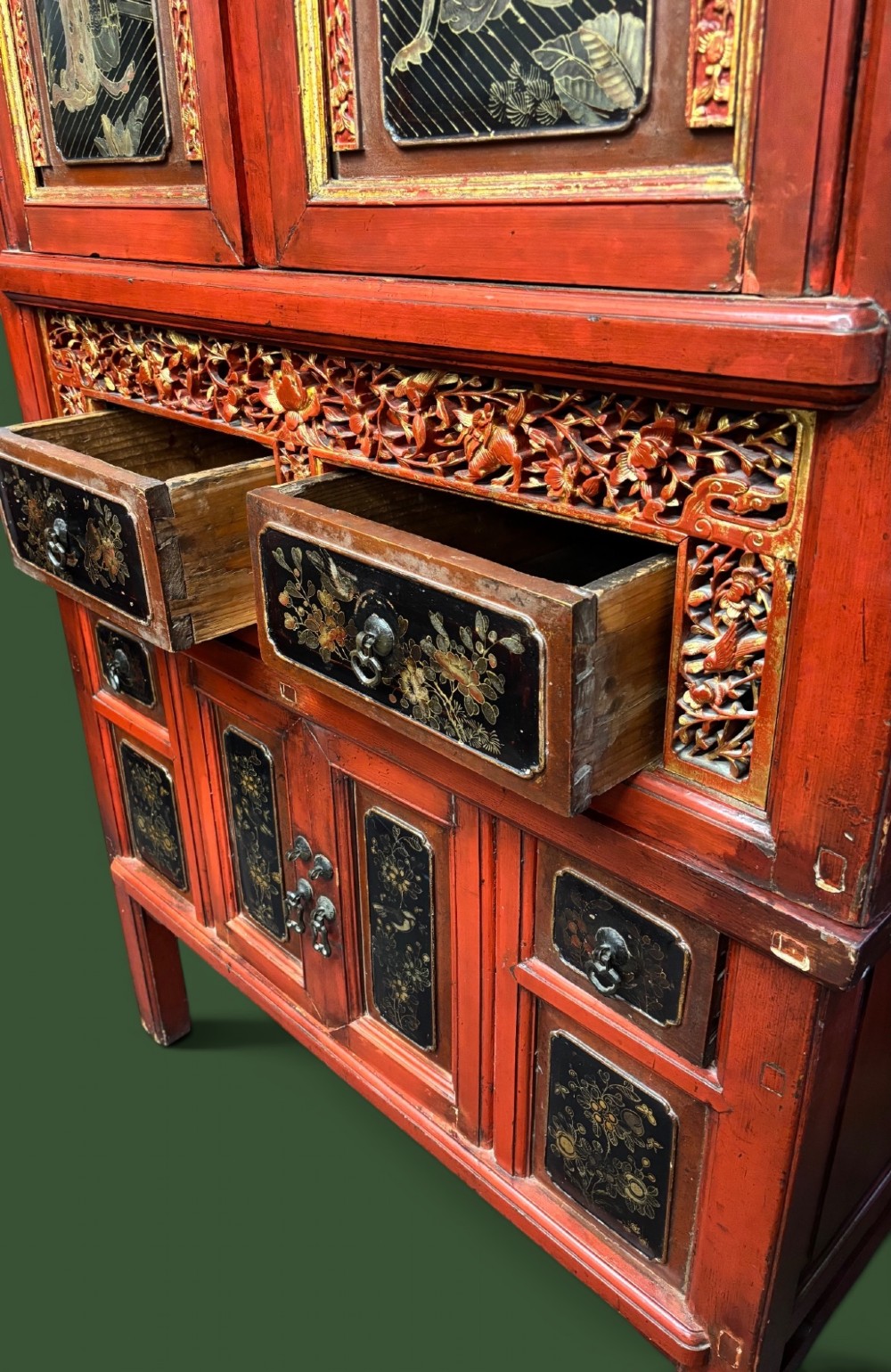 An antique Chinese tall wooden cabinet featuring red lacquered framing, gilded openwork wood carvings, black panels decorated with gold figures and floral motifs, and central brass hardware. View IV