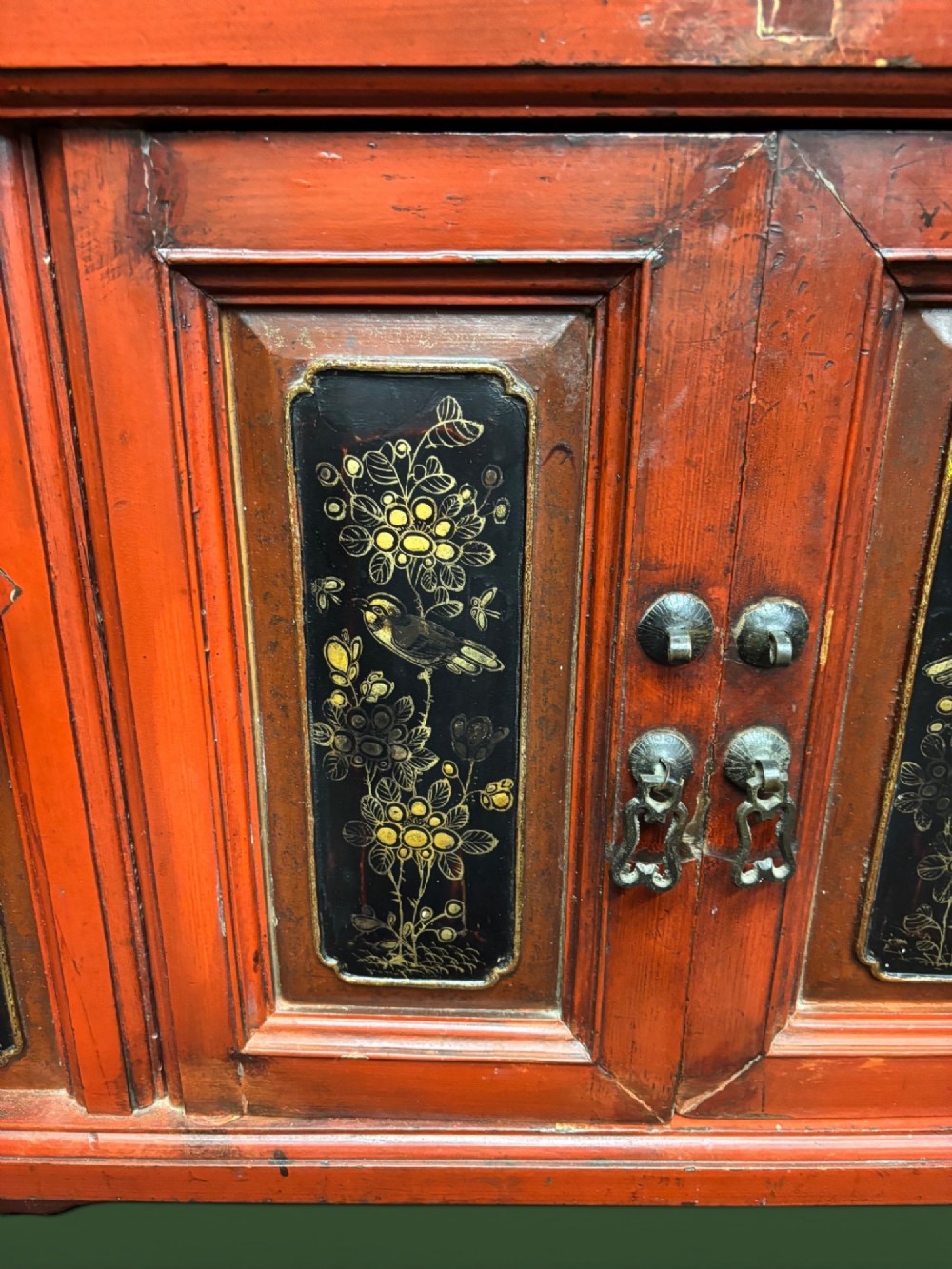 An antique Chinese tall wooden cabinet featuring red lacquered framing, gilded openwork wood carvings, black panels decorated with gold figures and floral motifs, and central brass hardware. View VI