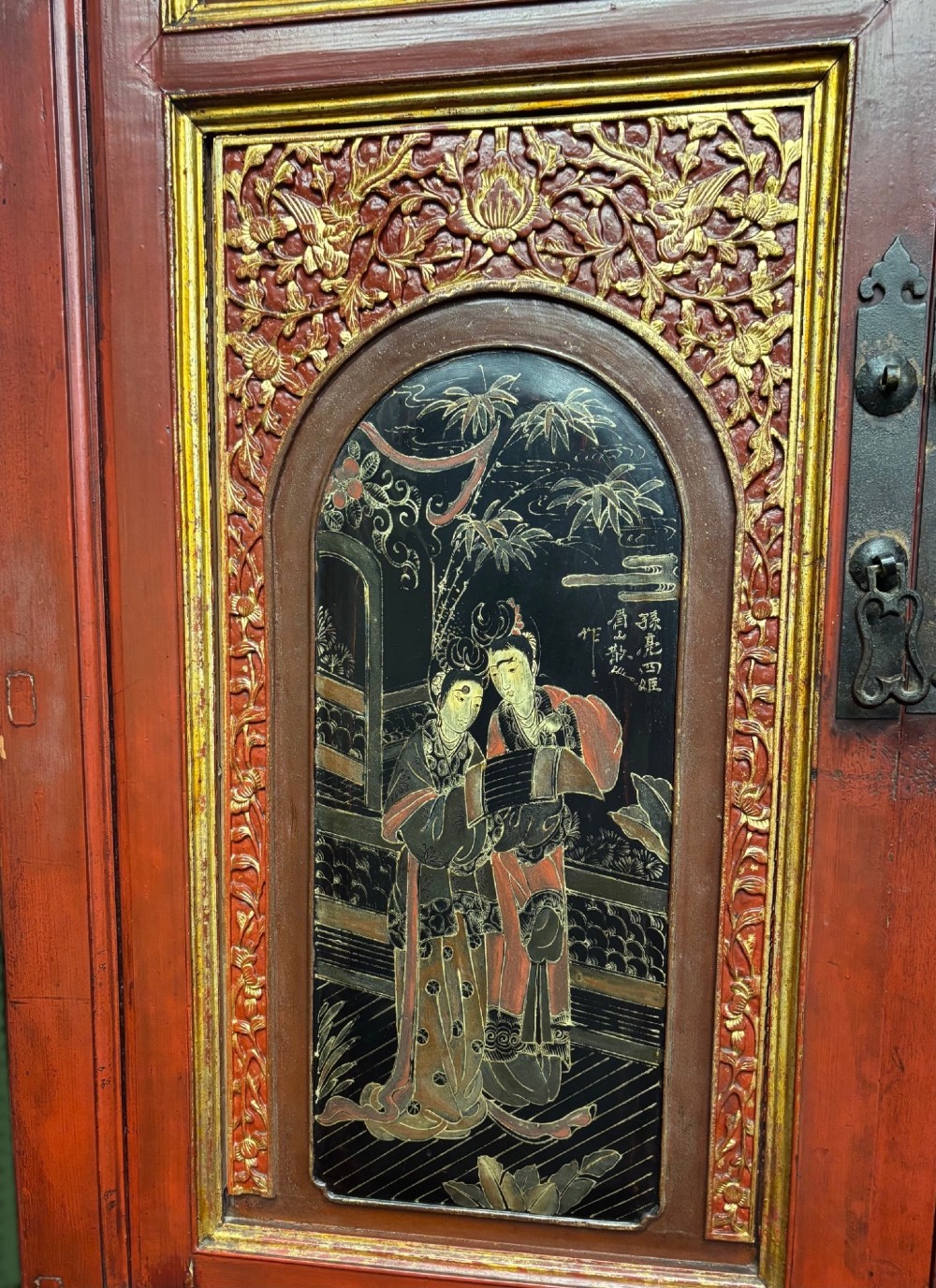 An antique Chinese tall wooden cabinet featuring red lacquered framing, gilded openwork wood carvings, black panels decorated with gold figures and floral motifs, and central brass hardware. View VII