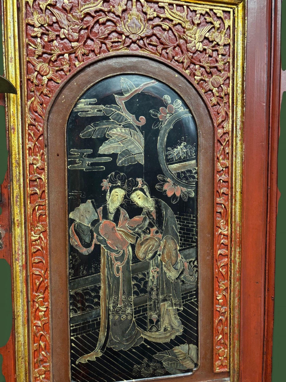 An antique Chinese tall wooden cabinet featuring red lacquered framing, gilded openwork wood carvings, black panels decorated with gold figures and floral motifs, and central brass hardware. View VIII