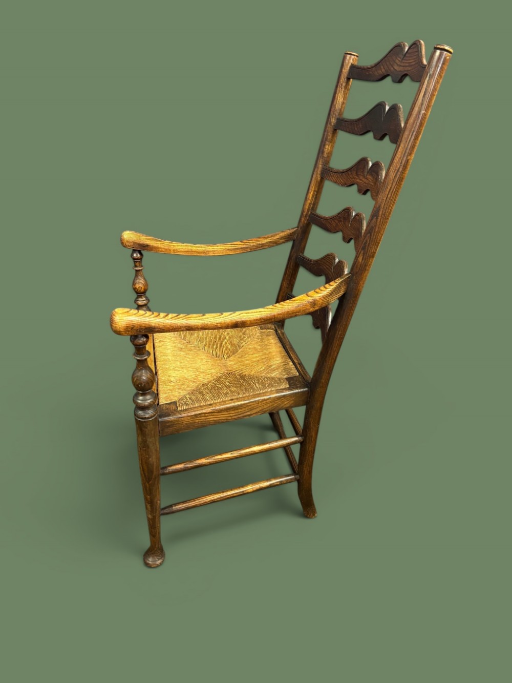An antique elm ladderback armchair featuring six shaped horizontal slats, curved armrests, and a woven rush seat. View II