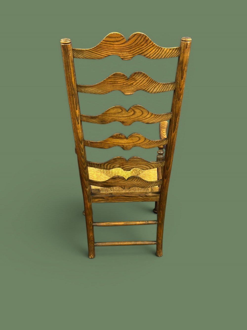 An antique elm ladderback armchair featuring six shaped horizontal slats, curved armrests, and a woven rush seat. View III