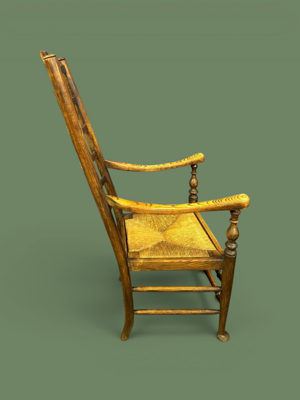 An antique elm ladderback armchair featuring six shaped horizontal slats, curved armrests, and a woven rush seat. View IV