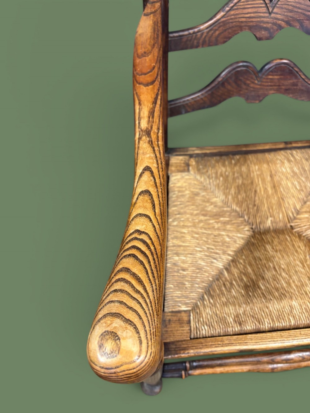 An antique elm ladderback armchair featuring six shaped horizontal slats, curved armrests, and a woven rush seat. View V