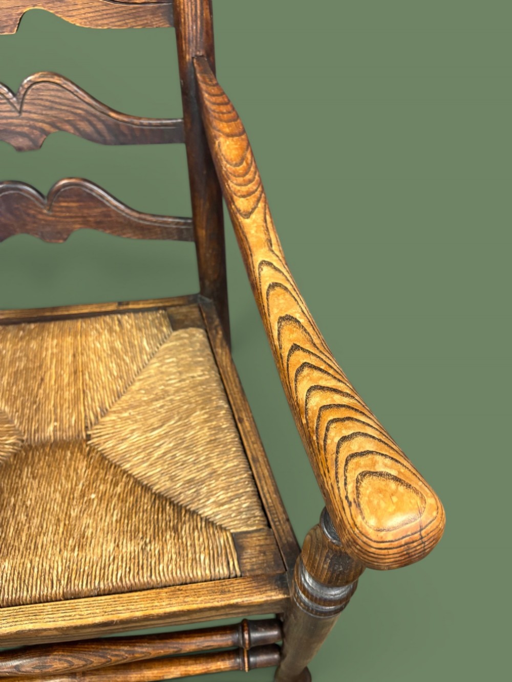 An antique elm ladderback armchair featuring six shaped horizontal slats, curved armrests, and a woven rush seat. View VI