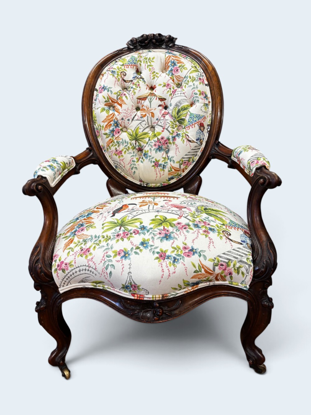 A Victorian carved mahogany armchair featuring an oval button-backed backrest, padded armrests, and botanical floral upholstery set on cabriole legs with brass castors.