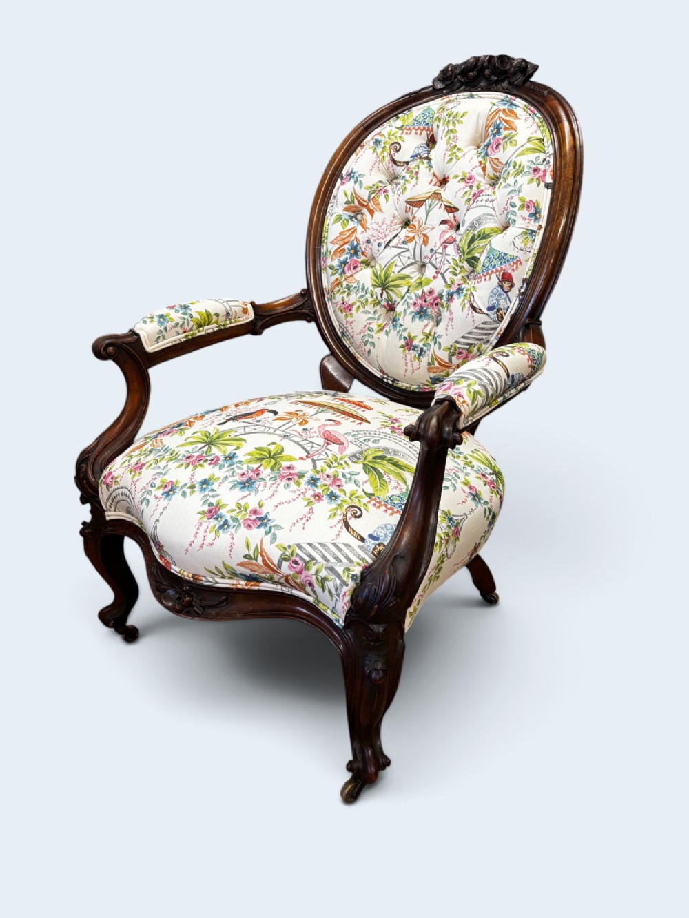 A Victorian carved mahogany armchair featuring an oval button-backed backrest, padded armrests, and botanical floral upholstery set on cabriole legs with brass castors. View II