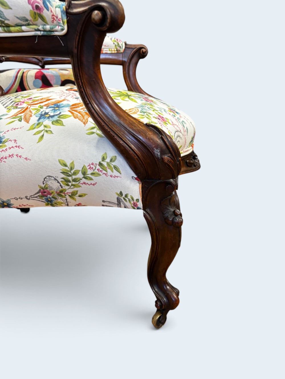 A Victorian carved mahogany armchair featuring an oval button-backed backrest, padded armrests, and botanical floral upholstery set on cabriole legs with brass castors. View IV