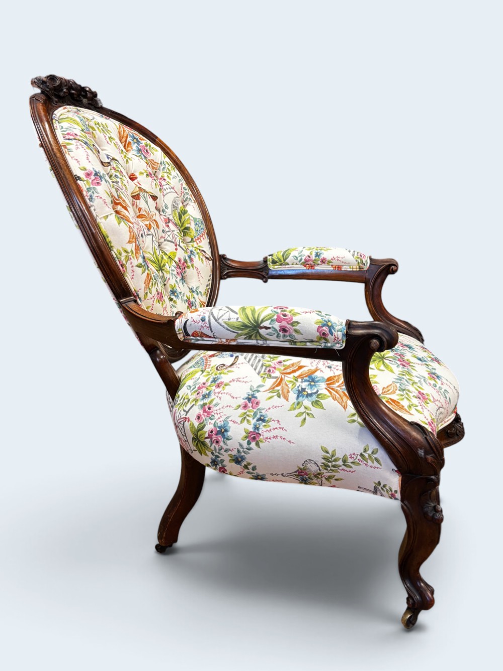 A Victorian carved mahogany armchair featuring an oval button-backed backrest, padded armrests, and botanical floral upholstery set on cabriole legs with brass castors. View V