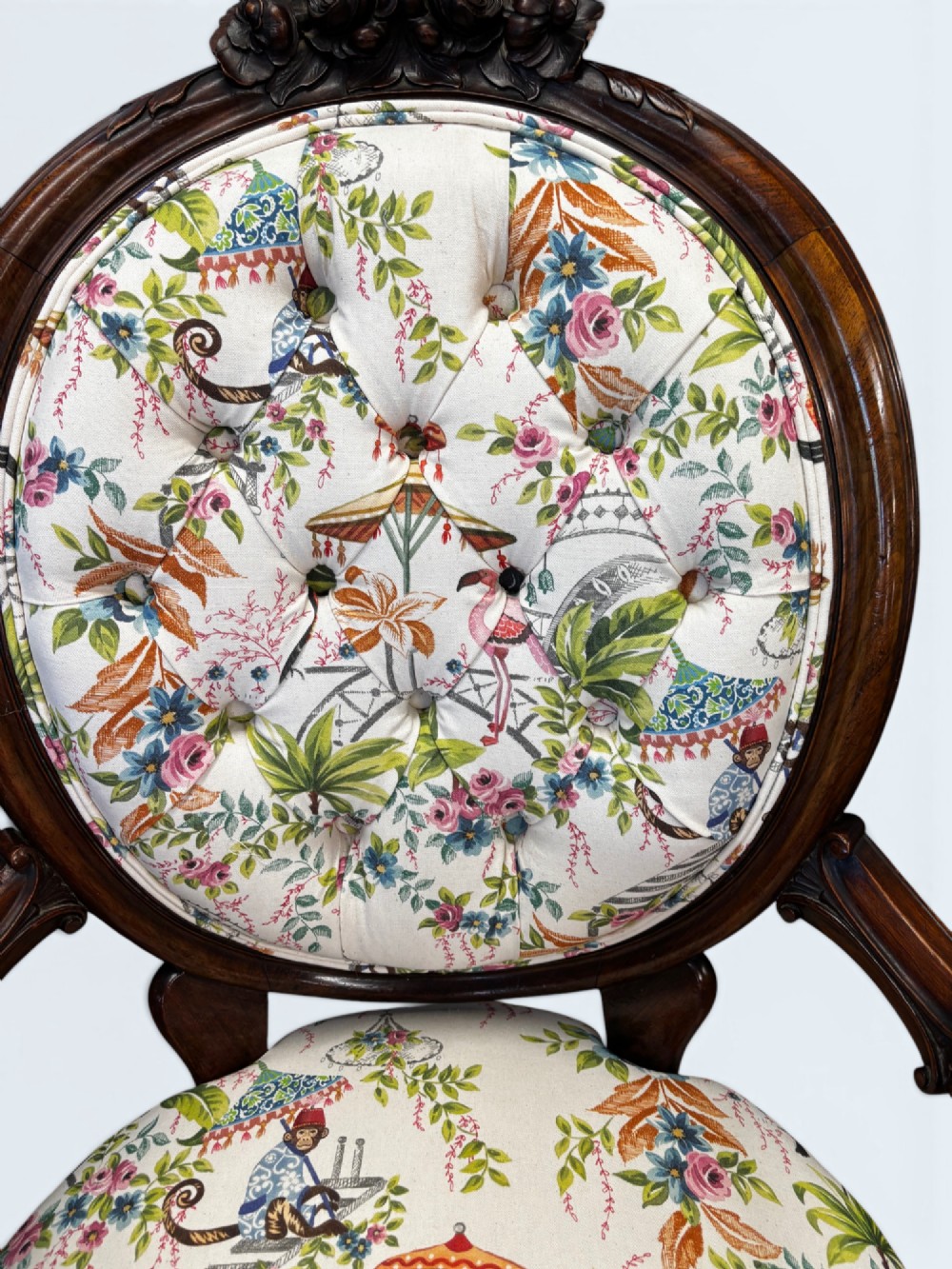 A Victorian carved mahogany armchair featuring an oval button-backed backrest, padded armrests, and botanical floral upholstery set on cabriole legs with brass castors. View VI