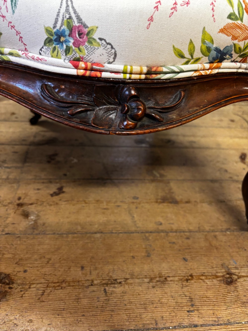A Victorian carved mahogany armchair featuring an oval button-backed backrest, padded armrests, and botanical floral upholstery set on cabriole legs with brass castors. View VIII