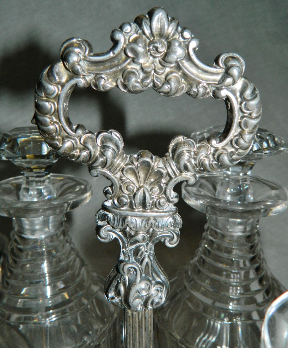 A Victorian silver-plated decanter stand featuring a central foliate handle and four matching cut-glass decanters with faceted stoppers. View V