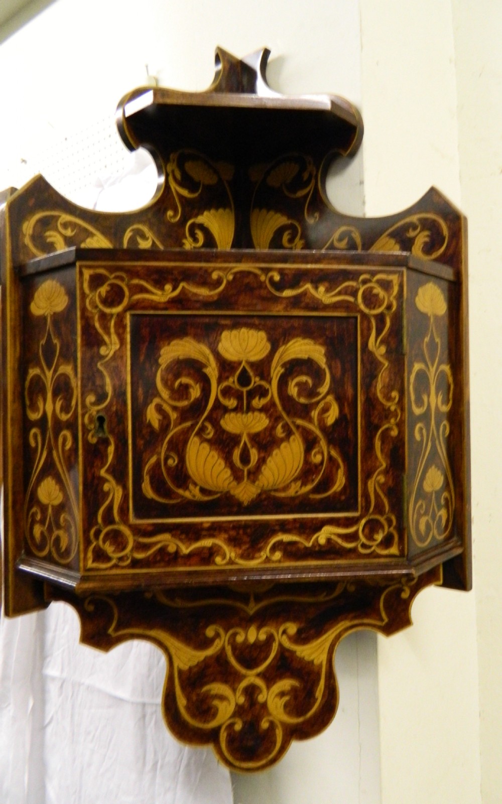 A dark wood hanging wall cabinet decorated with intricate floral light-wood marquetry inlay, featuring a single door and a shaped decorative gallery.