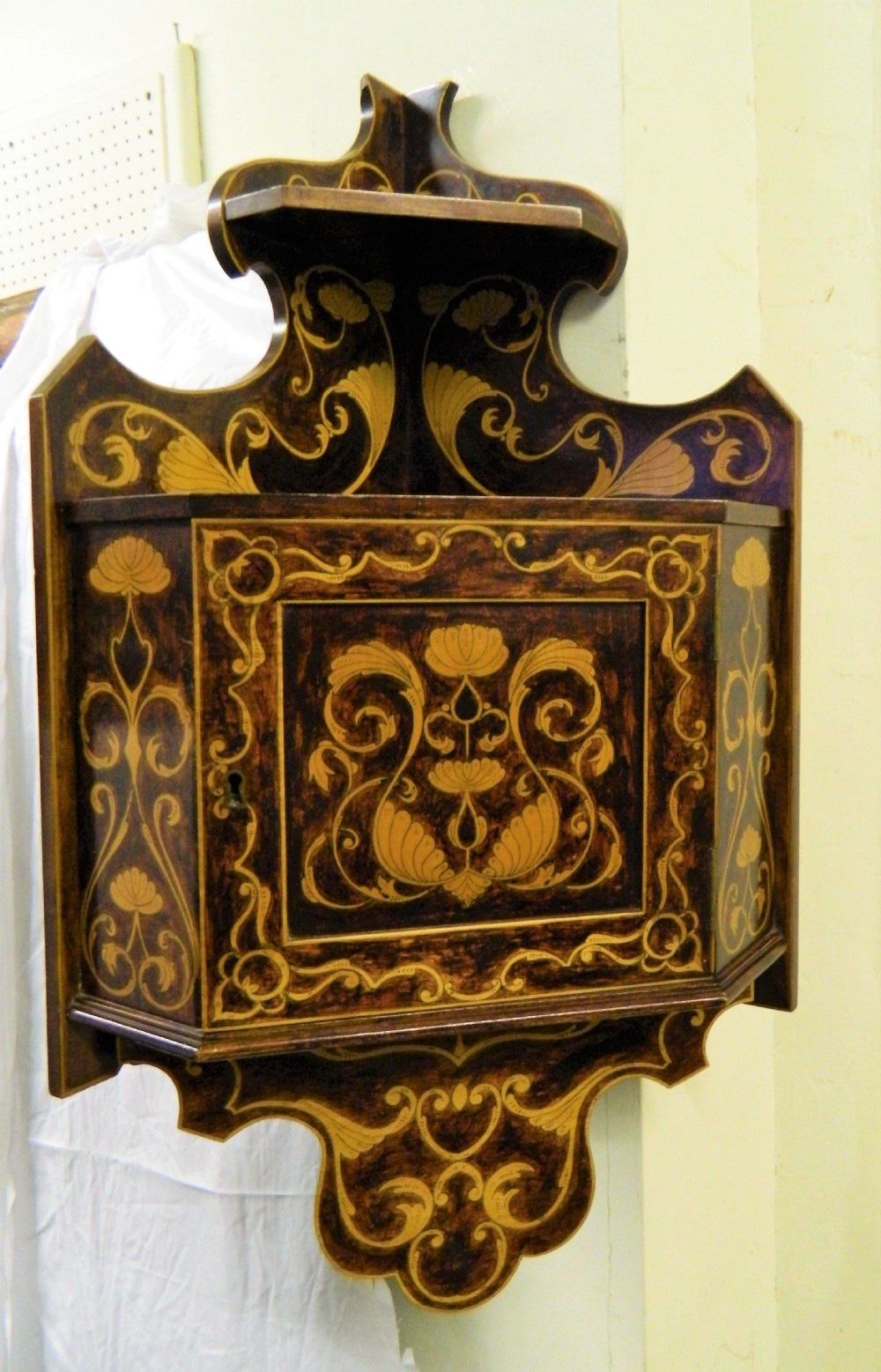 A dark wood hanging wall cabinet decorated with intricate floral light-wood marquetry inlay, featuring a single door and a shaped decorative gallery. View VI