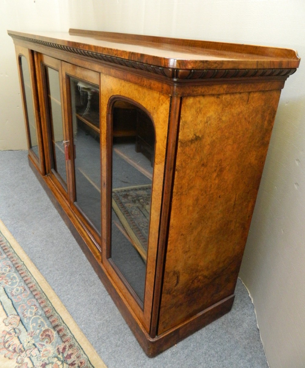 A large Victorian burr walnut bookcase featuring four glass-panelled doors, detailed wood grain patterning, and a solid plinth base. View VI