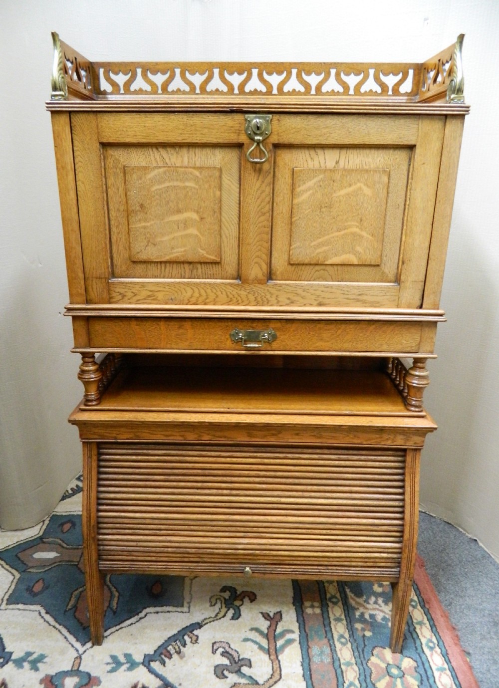 An Arts and Crafts golden oak bureau featuring a pierced heart gallery top, a fall-front writing section, a single drawer, an open spindle shelf, and a lower tambour front cabinet.