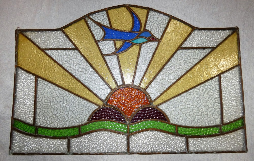 An Art Deco arched leaded stained glass panel depicting a blue bird flying above a sunburst motif over green hills.