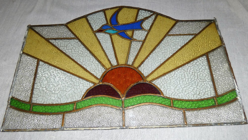 An Art Deco arched leaded stained glass panel depicting a blue bird flying above a sunburst motif over green hills. View V