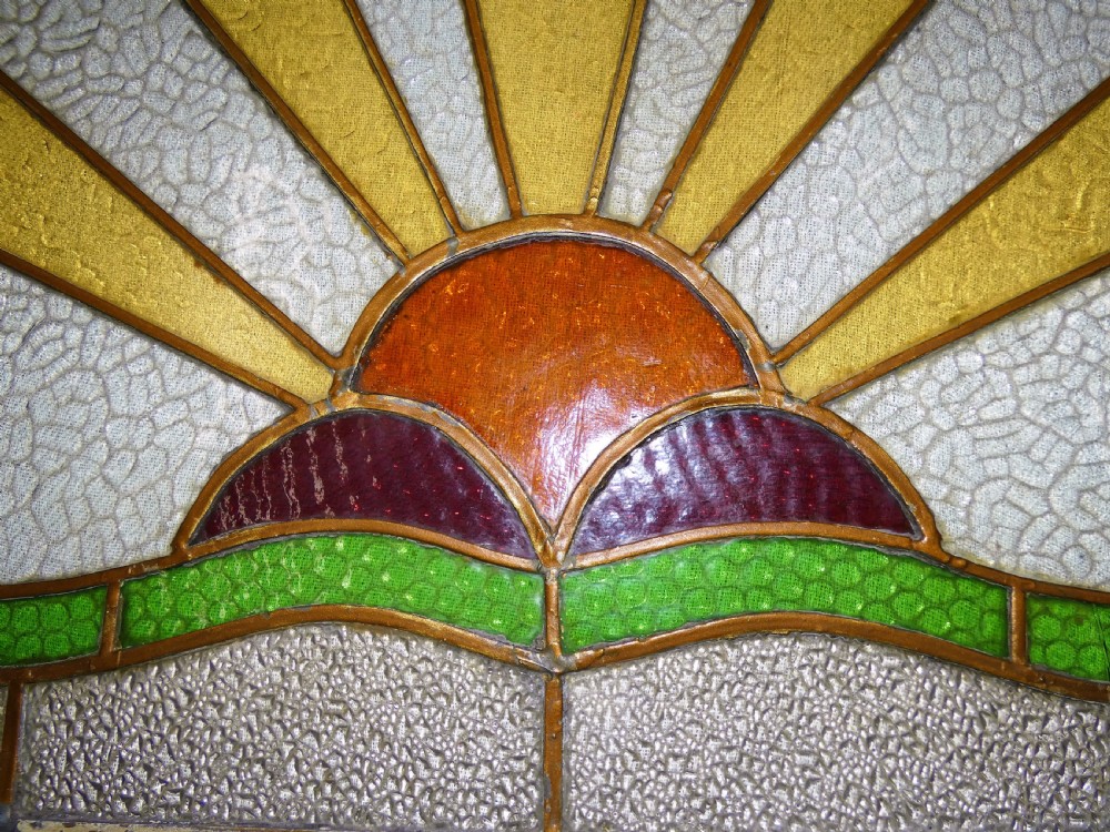 An Art Deco arched leaded stained glass panel depicting a blue bird flying above a sunburst motif over green hills. View VI