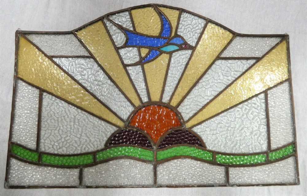 An Art Deco arched leaded stained glass panel depicting a blue bird flying above a sunburst motif over green hills. View VII