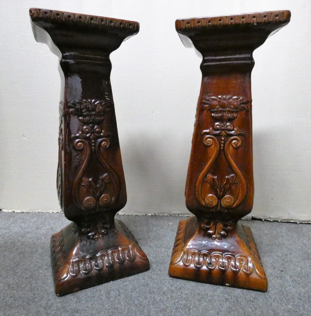 A pair of Arts and Crafts warm brown glazed earthenware plant stands featuring moulded relief scrollwork and square bases.
