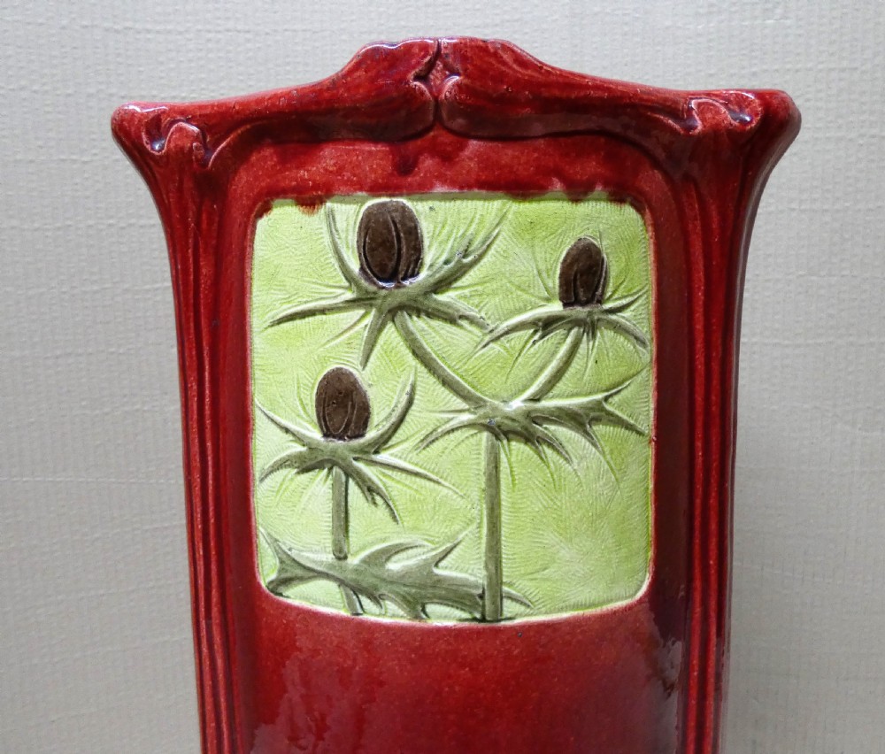 A tall cylindrical ceramic stick stand finished in a deep red glaze featuring an inset green relief panel with three thistle motifs. View II