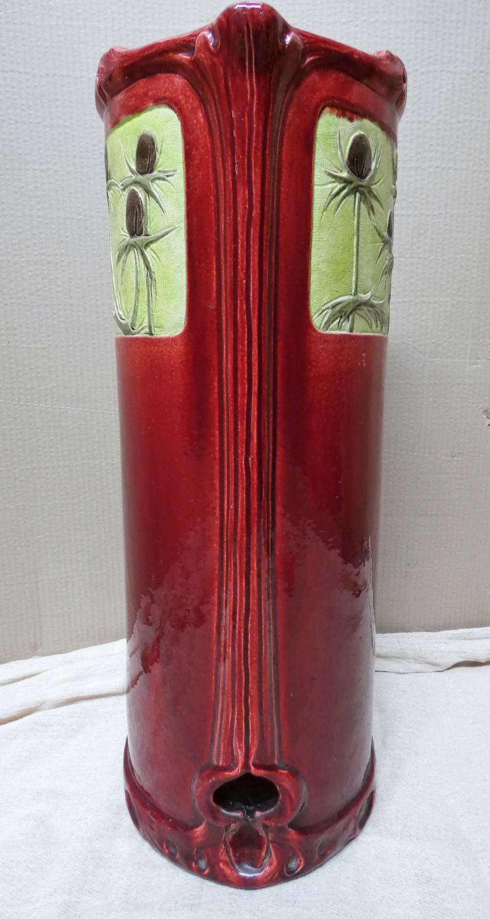 A tall cylindrical ceramic stick stand finished in a deep red glaze featuring an inset green relief panel with three thistle motifs. View III