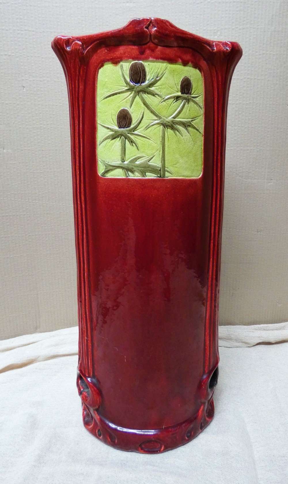 A tall cylindrical ceramic stick stand finished in a deep red glaze featuring an inset green relief panel with three thistle motifs. View V