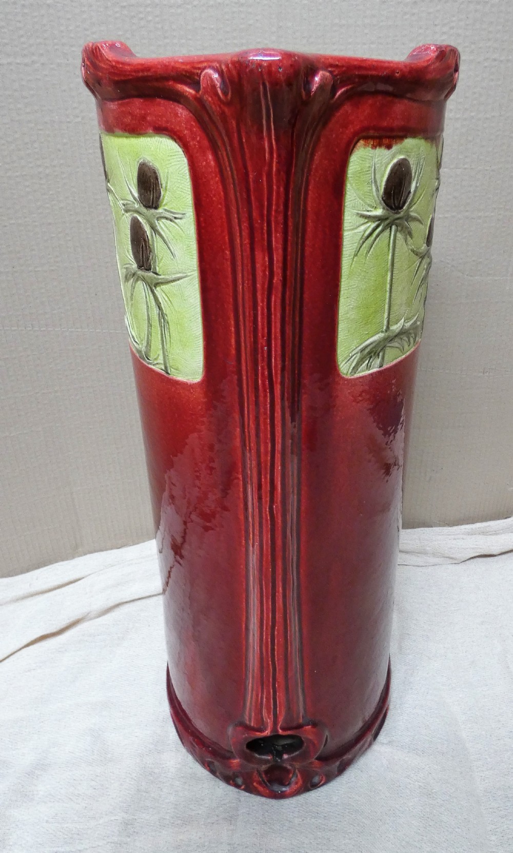 A tall cylindrical ceramic stick stand finished in a deep red glaze featuring an inset green relief panel with three thistle motifs. View VII
