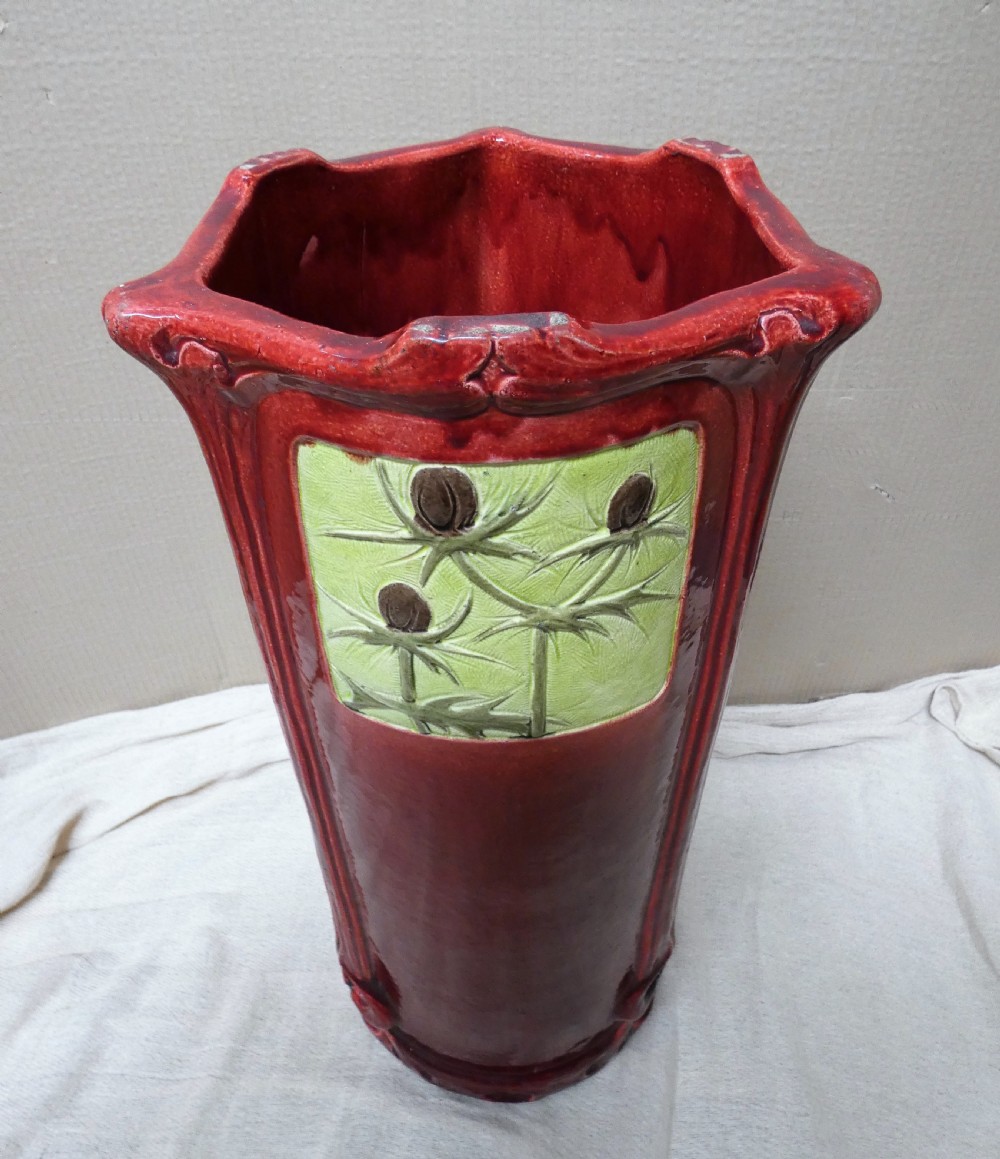 A tall cylindrical ceramic stick stand finished in a deep red glaze featuring an inset green relief panel with three thistle motifs. View VIII