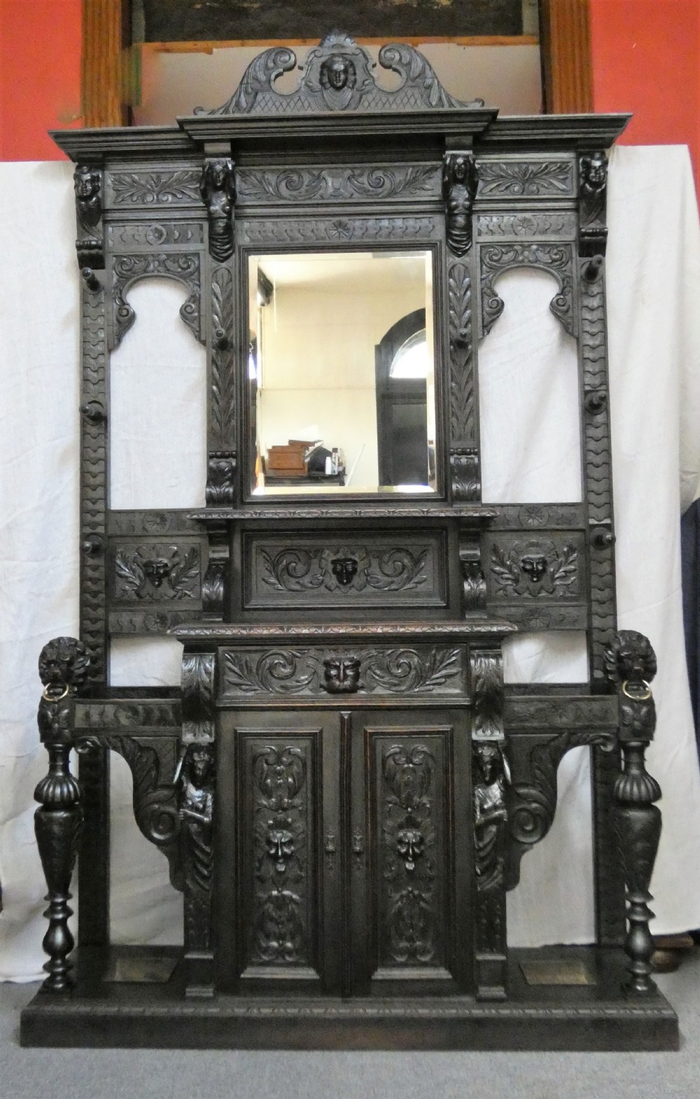 A tall dark carved oak hall stand featuring a central rectangular mirror, carved mask motifs, side coat pegs, an integrated cupboard, and twin umbrella stands.
