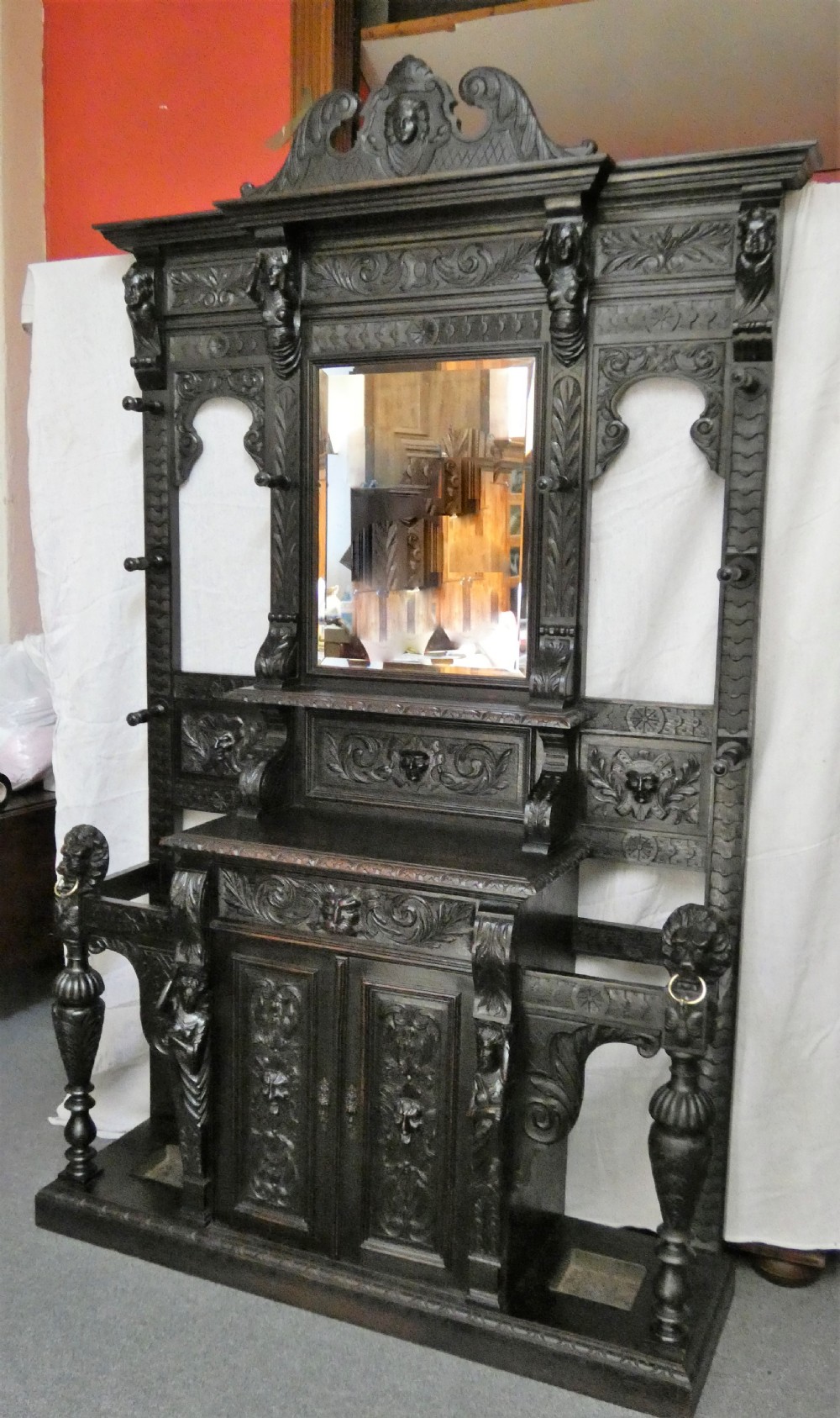 A tall dark carved oak hall stand featuring a central rectangular mirror, carved mask motifs, side coat pegs, an integrated cupboard, and twin umbrella stands. View II