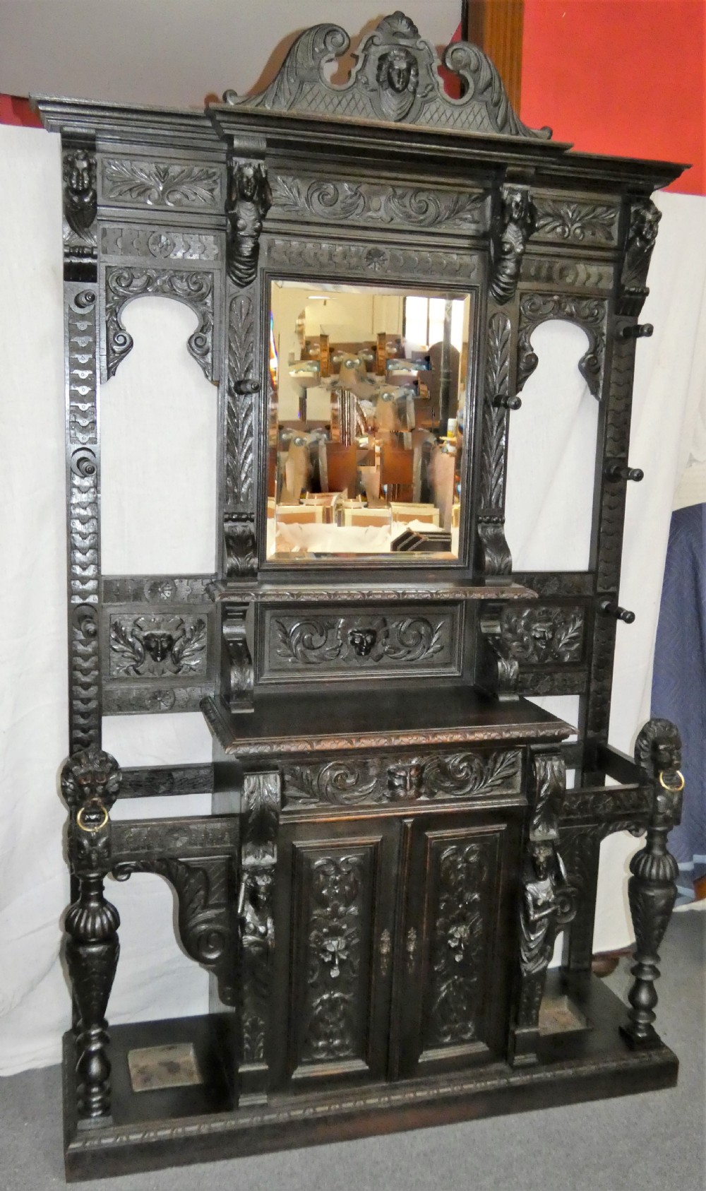 A tall dark carved oak hall stand featuring a central rectangular mirror, carved mask motifs, side coat pegs, an integrated cupboard, and twin umbrella stands. View III