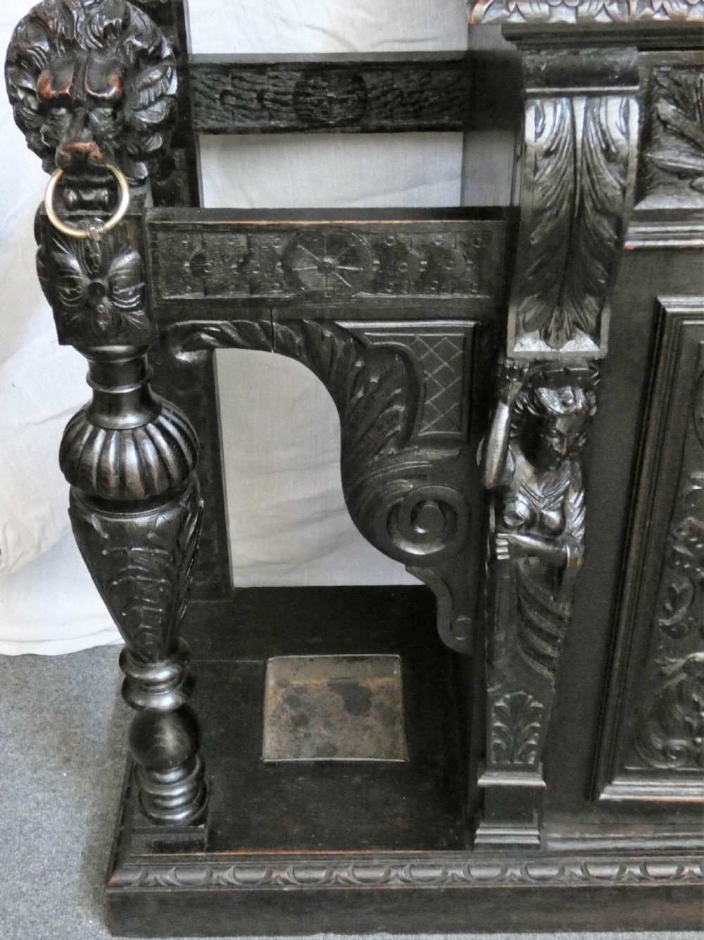 A tall dark carved oak hall stand featuring a central rectangular mirror, carved mask motifs, side coat pegs, an integrated cupboard, and twin umbrella stands. View V