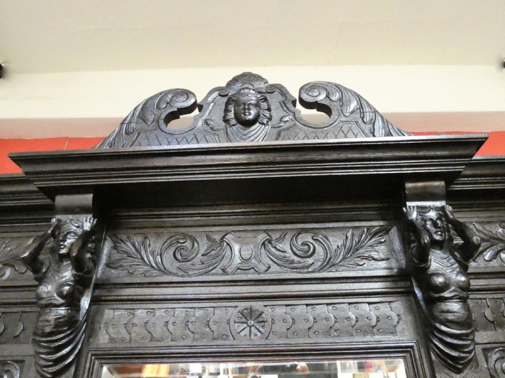 A tall dark carved oak hall stand featuring a central rectangular mirror, carved mask motifs, side coat pegs, an integrated cupboard, and twin umbrella stands. View VI