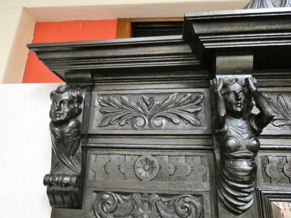 A tall dark carved oak hall stand featuring a central rectangular mirror, carved mask motifs, side coat pegs, an integrated cupboard, and twin umbrella stands. View VII