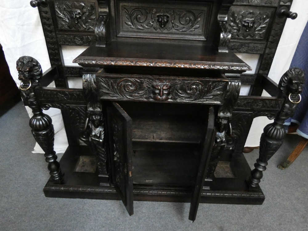 A tall dark carved oak hall stand featuring a central rectangular mirror, carved mask motifs, side coat pegs, an integrated cupboard, and twin umbrella stands. View VIII