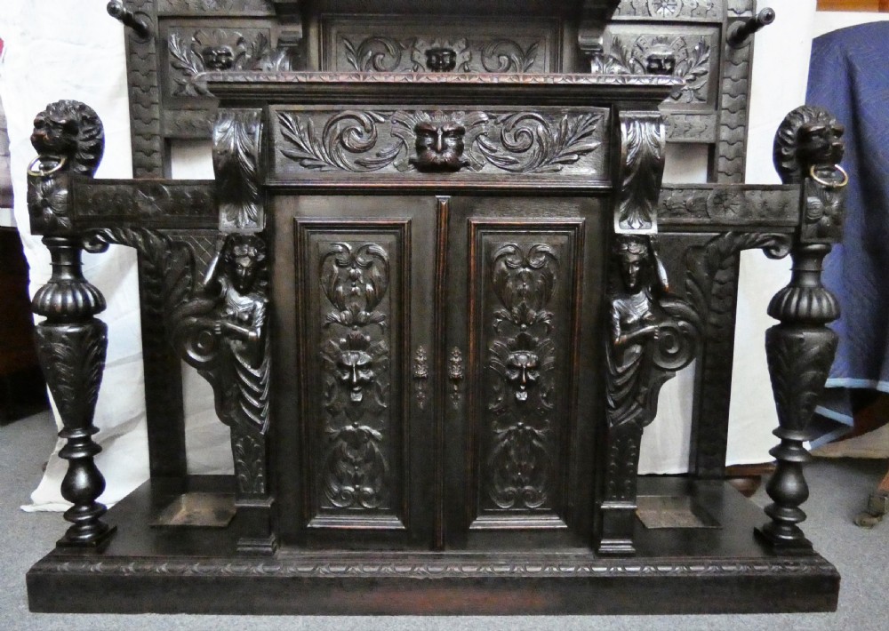 A tall dark carved oak hall stand featuring a central rectangular mirror, carved mask motifs, side coat pegs, an integrated cupboard, and twin umbrella stands. View IX