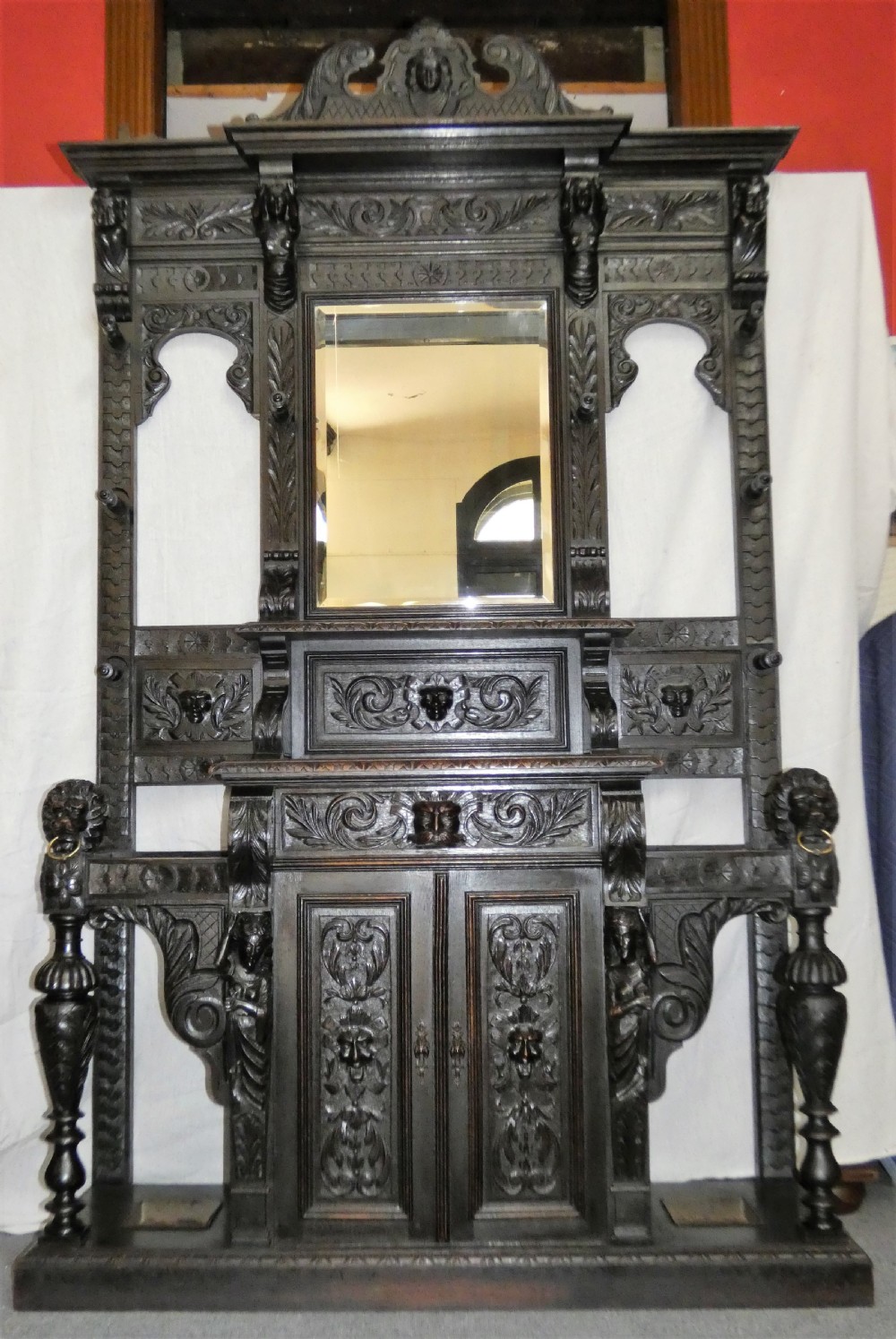 A tall dark carved oak hall stand featuring a central rectangular mirror, carved mask motifs, side coat pegs, an integrated cupboard, and twin umbrella stands. View X