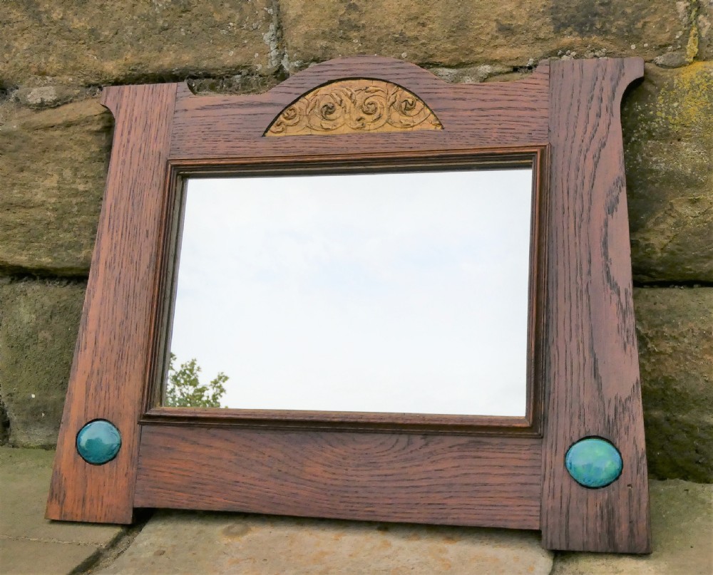 An Arts and Crafts style dark oak wall mirror featuring a carved arched crest and two round turquoise ceramic inlays in the lower corners.