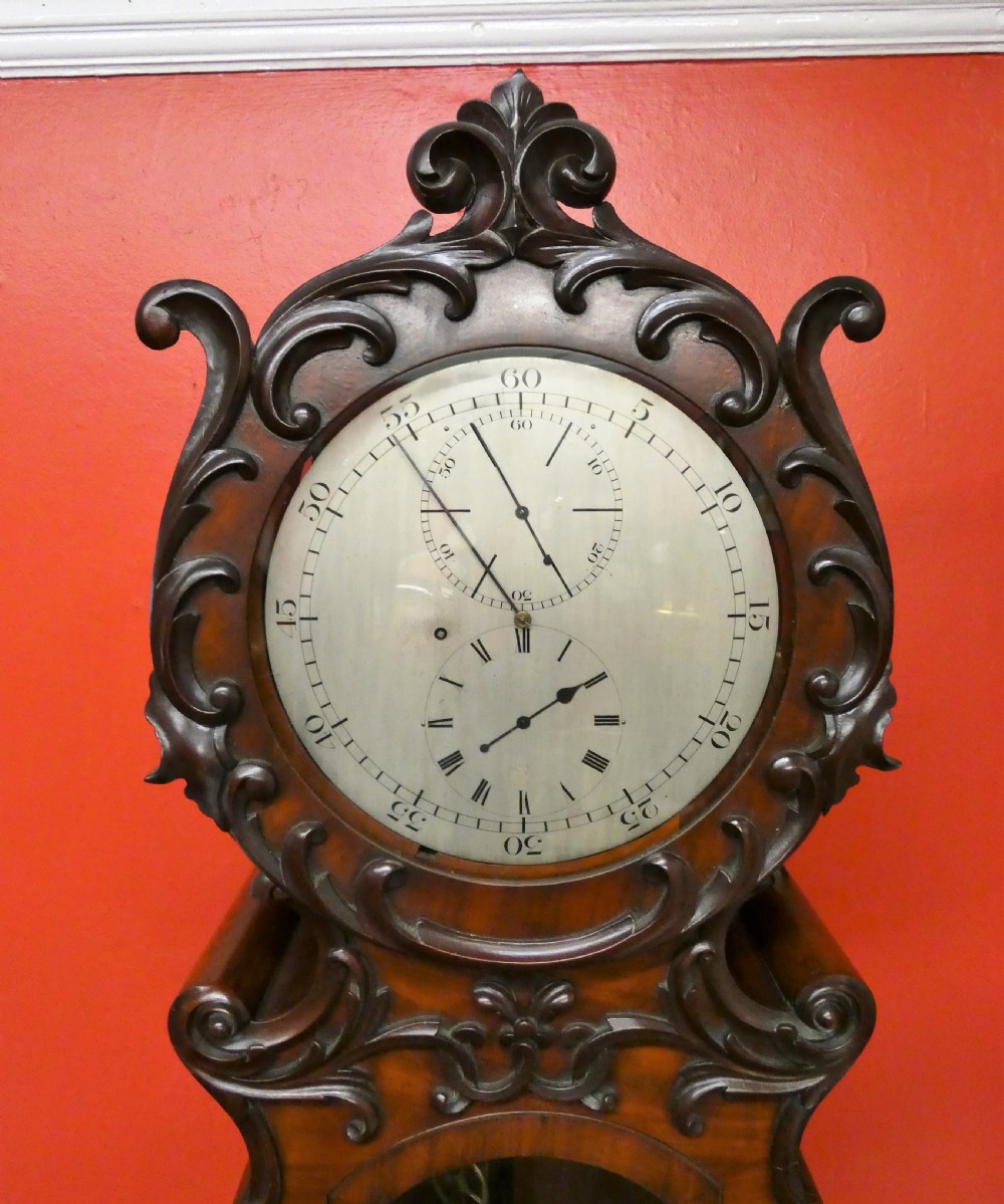 An antique carved dark mahogany regulator longcase clock featuring a circular dial, glass trunk door displaying pendulum weights, and a carved pedestal base. View II
