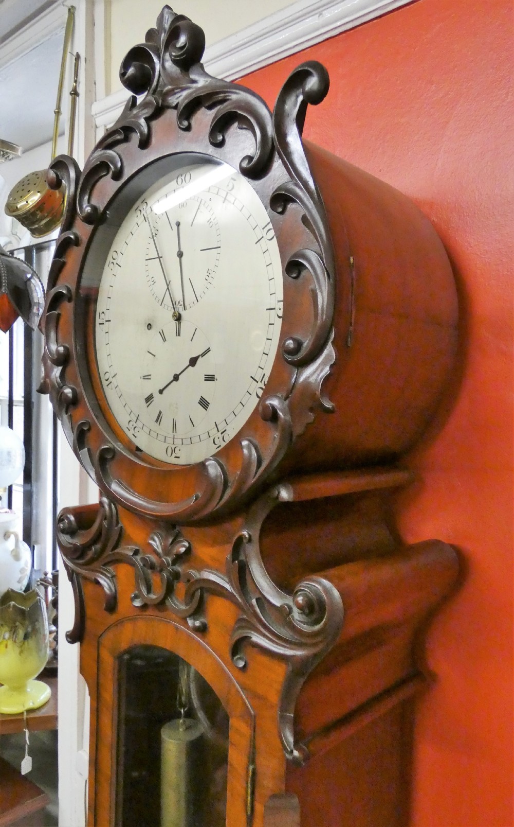 An antique carved dark mahogany regulator longcase clock featuring a circular dial, glass trunk door displaying pendulum weights, and a carved pedestal base. View VI