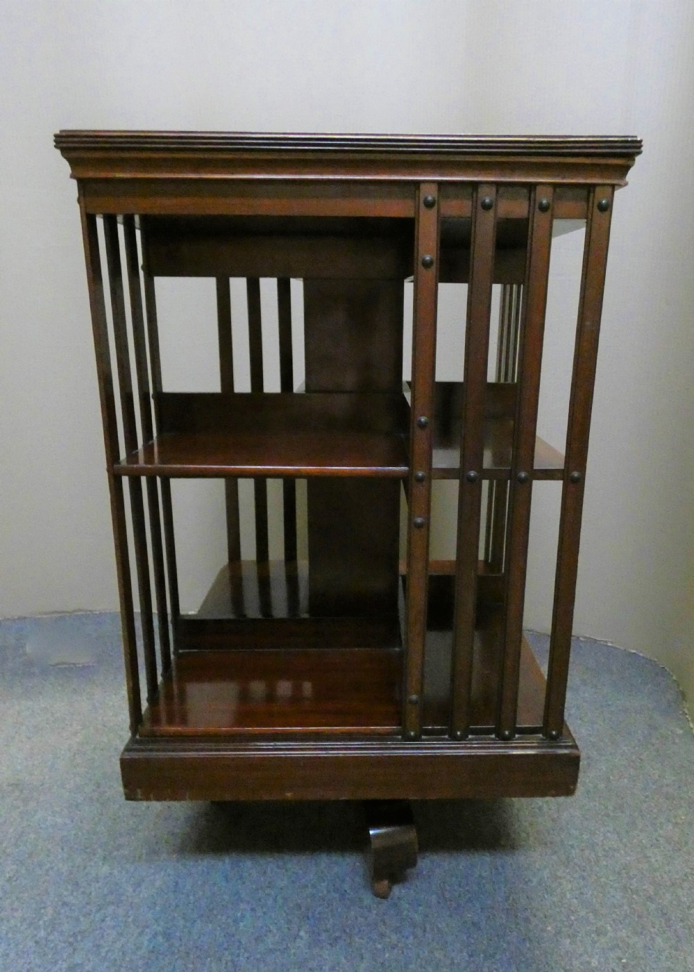 A dark polished mahogany revolving bookcase featuring vertical slatted sides with metal stud detail, multiple storage tiers, and a rotating base on castors.