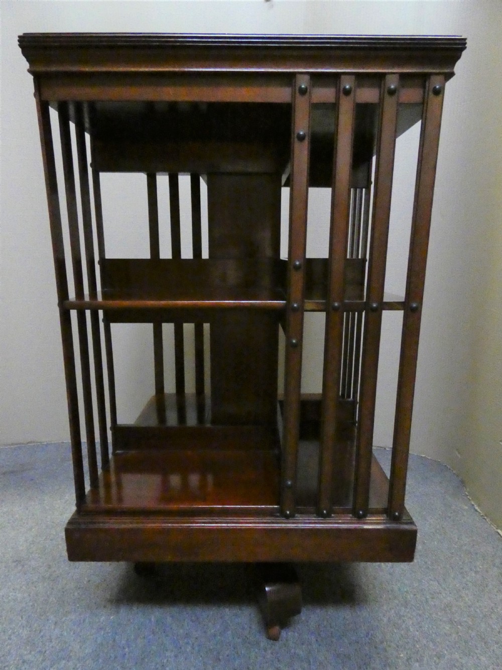 A dark polished mahogany revolving bookcase featuring vertical slatted sides with metal stud detail, multiple storage tiers, and a rotating base on castors. View II