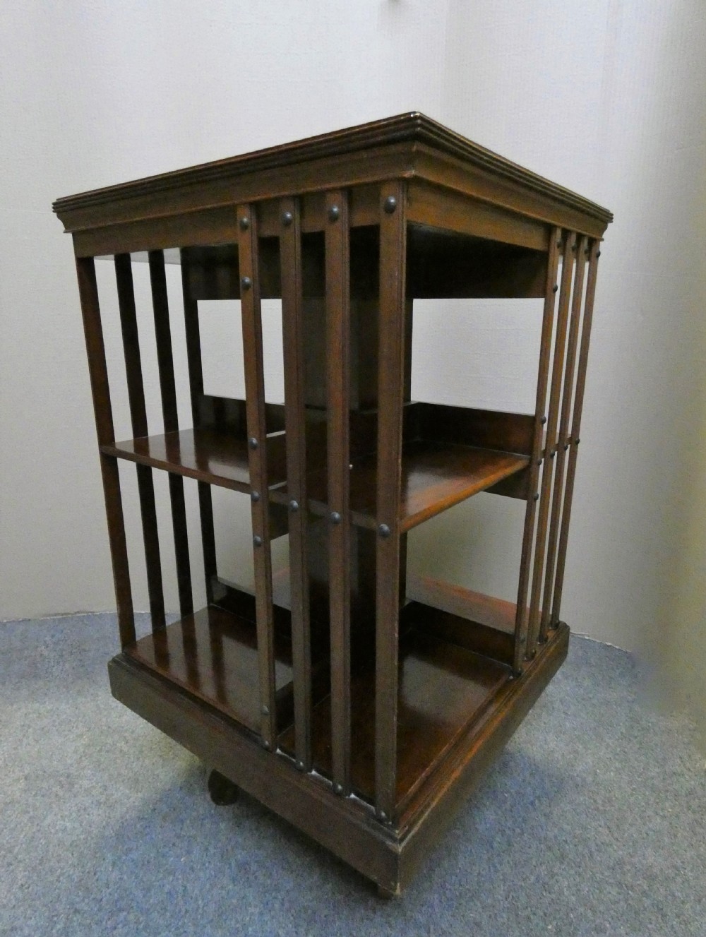 A dark polished mahogany revolving bookcase featuring vertical slatted sides with metal stud detail, multiple storage tiers, and a rotating base on castors. View III