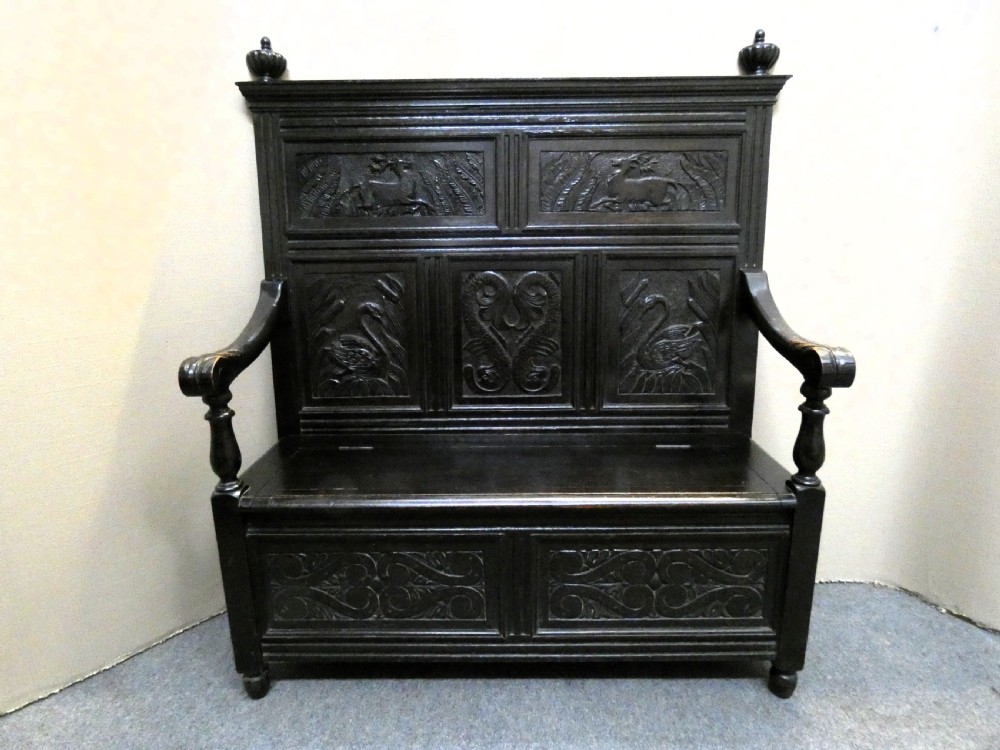A dark carved oak high-back settle featuring relief panel carvings of swans and stags, turned arm supports, and a hinged box seat.